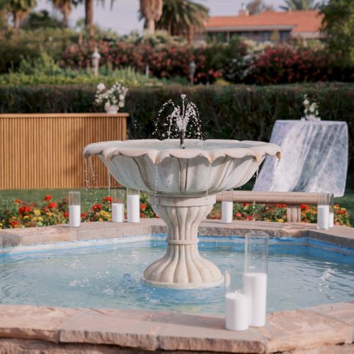 A stone fountain splashes into a swimming pool, with candles, lush flowers, and tall palm trees in a landscaped garden behind it.